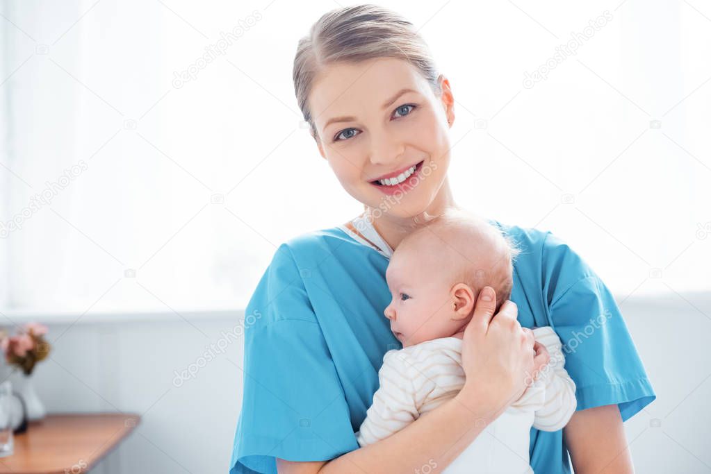 happy young mother holding newborn baby and smiling at camera in hospital room