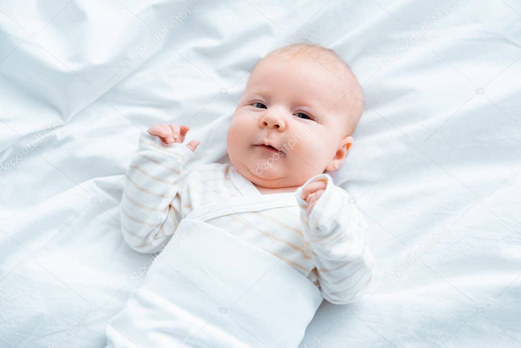 top view of adorable baby looking at camera while lying on white bedding 