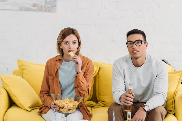 異人種間のカップル楽しむ軽食や黄色のソファーに座りながら飲み物 — ストック写真