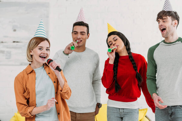 multiethnic multicultural friends in colorful paper hats having fun at home party while singing karaoke 