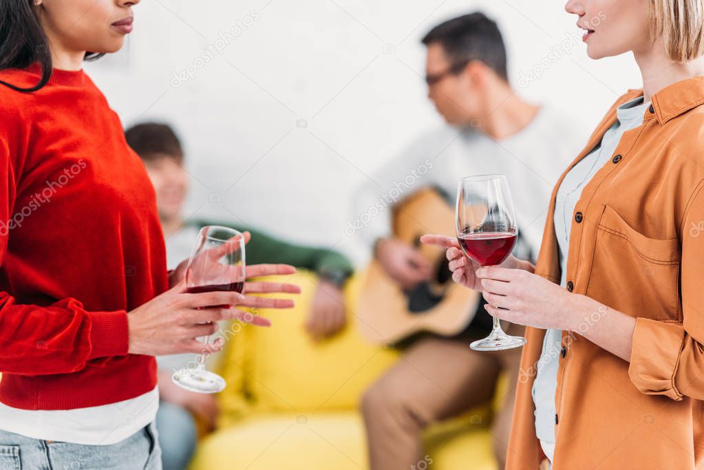multicultural girls holding wine glasses and talking, men sitting on yellow sofa on background 