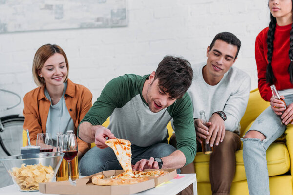 young man taking piece of pizza while multicultural friends enjoying drinks