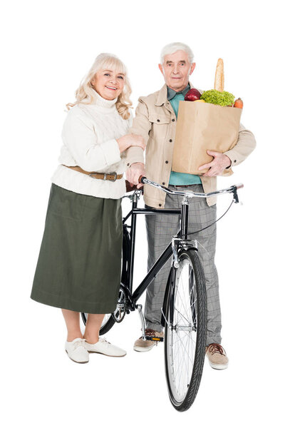 happy wife standing with retired husband holding bike and paper bag isolated on white