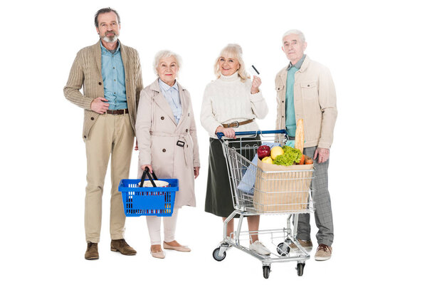 cheerful senior friends standing with basket and shopping cart isolated on white