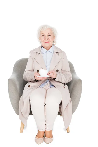 茶白で隔離のカップと肘掛け椅子に座っている陽気な年配の女性 — ストック写真