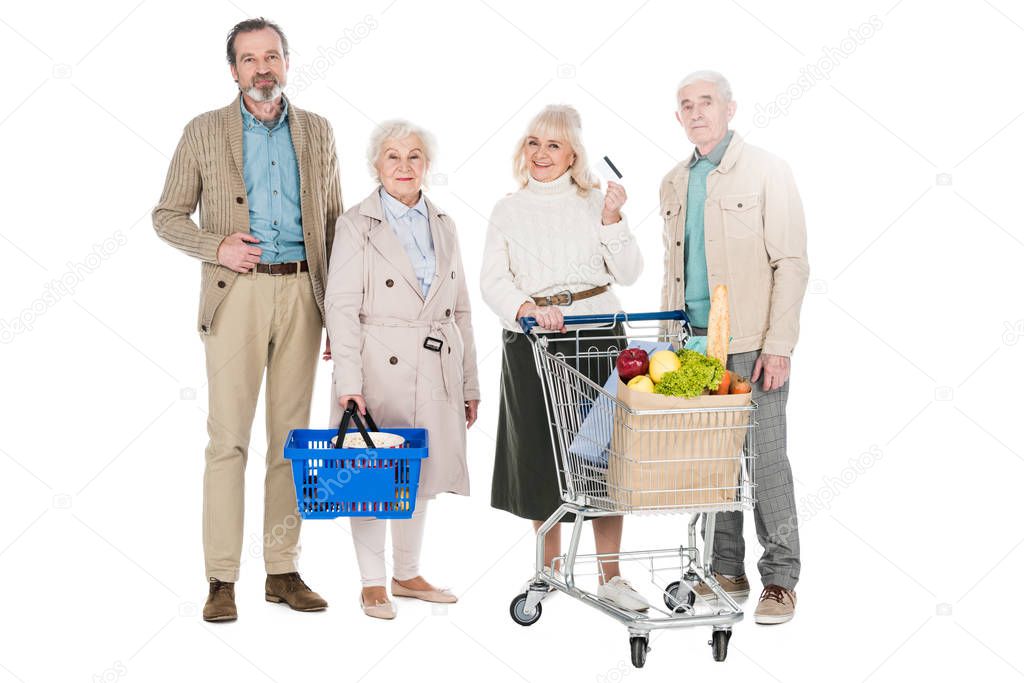 cheerful senior friends standing with basket and shopping cart isolated on white