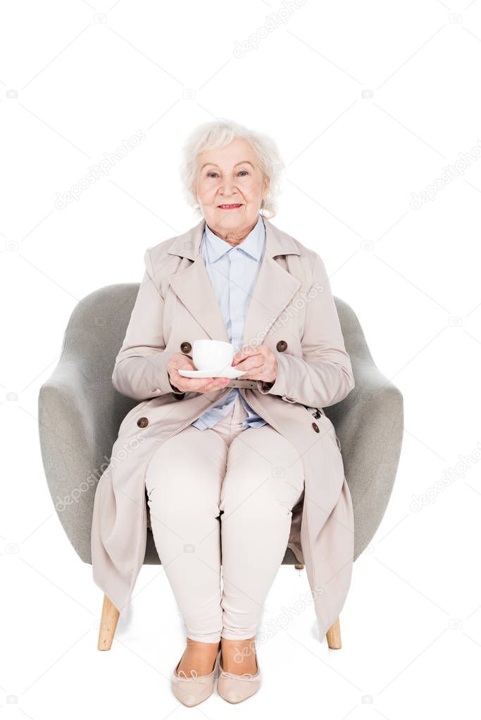cheerful senior woman sitting in armchair with cup of tea isolated on white