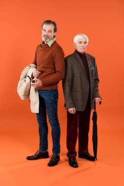 trendy retired man standing with jacket in hands near friend with umbrella on orange background clipart