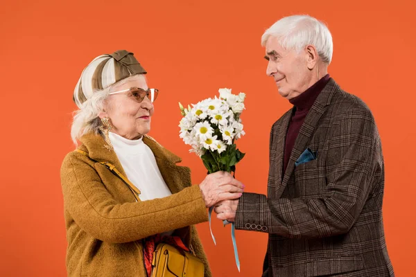 happy senior man giving flowers to stylish retired wife isolated on orange