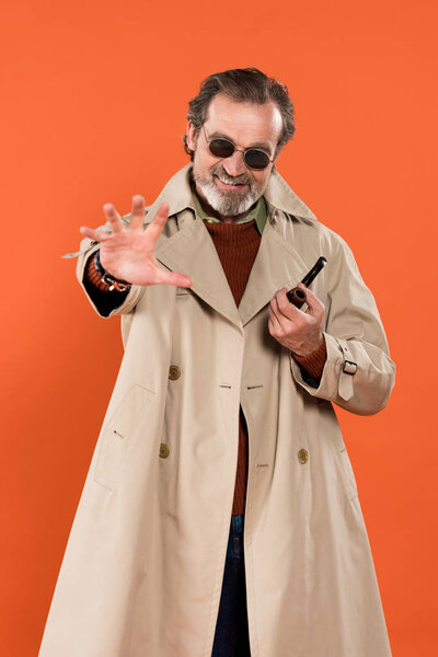 trendy senior man holding smoking pipe while standing isolated on orange 