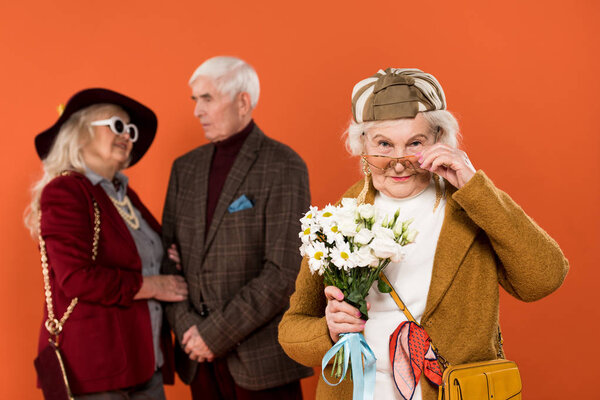 селективное внимание стильной пожилой женщины в солнцезащитных очках, держащей букет с парой пенсионеров на фоне изолированного оранжевого
 