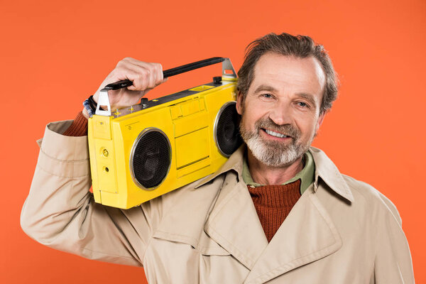 cheerful senior man holding yellow boombox isolated on orange