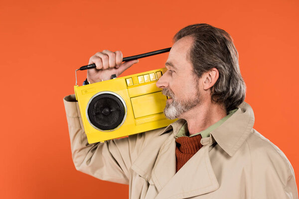 cheerful retired man looking at yellow boombox isolated on orange