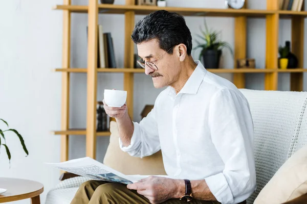Emeryturze Człowiek Okularach Trzymając Kubek Kawą Czytanie Gazety Domu — Zdjęcie stockowe