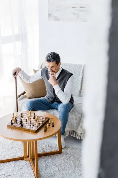 pensive retired man with mustache looking at chess board while holding walking cane at home