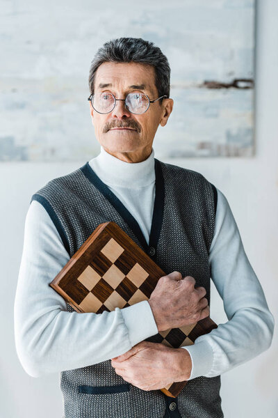 pensive senior man holding chess board at home