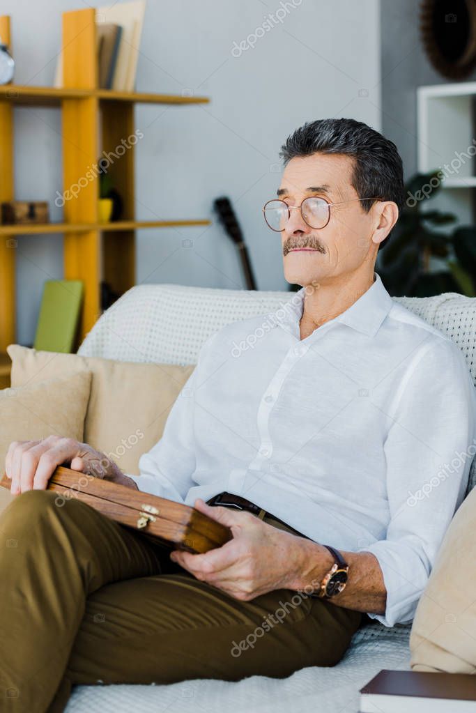 Pensioner in glasses holding chess board while sitting on sofa