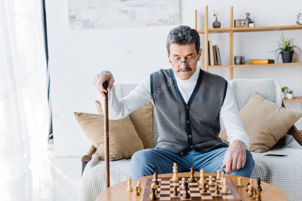 retired man with mustache looking at chess board while holding walking cane at home
