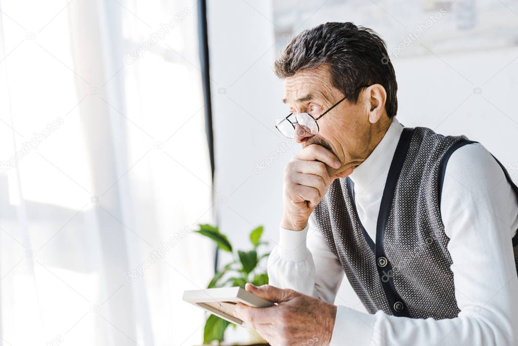 pensive retired man in glasses holding photo frame while sitting at home