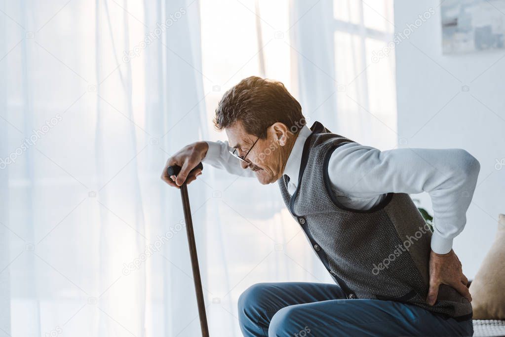 senior man with walking cane holding back while standing up from sofa