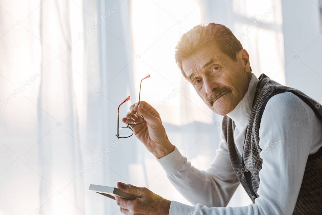 sad pensioner holding glasses and looking at camera while sitting at home