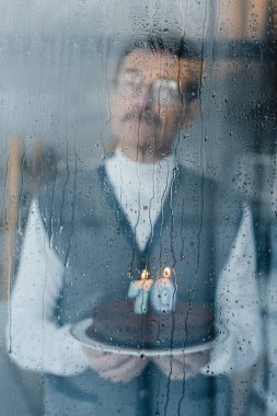 selective focus of window with raindrops with pensive senior man holding birthday cake on background clipart