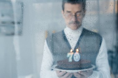 selective focus of window with raindrops with sad senior man holding birthday cake on background clipart