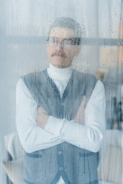 pensive retired man standing with crossed arms near window 