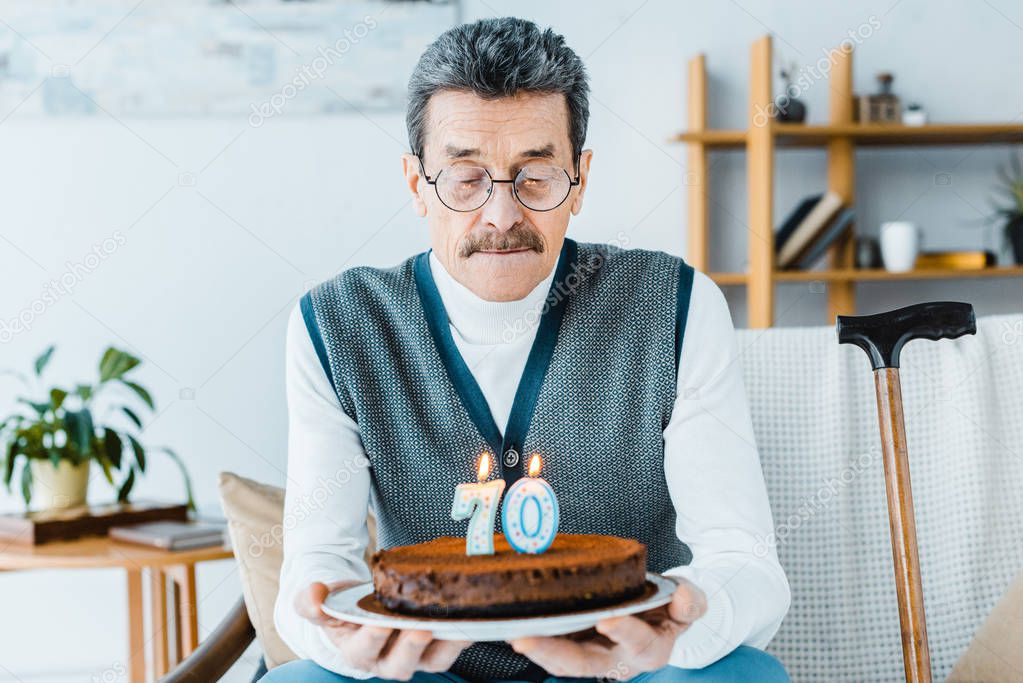 sad senior man holding birthday cake while sitting on sofa a in living room 