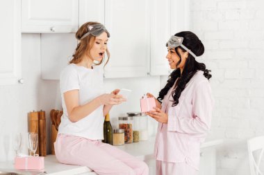 uyku maskesi ve Afro-Amerikan kız mutfakta hediye kutusu tutarak iken smartphone kullanarak Pijama içinde güzel kız
