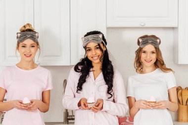 beautiful multiethnic girls in sleeping masks holding cupcakes and looking at camera during pajama party clipart