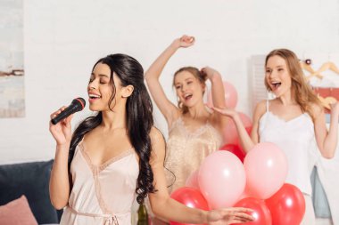beautiful multiethnic girls in nightwear singing karaoke at pajama party in bedroom clipart