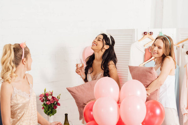 beautiful happy multiethnic girls in nightwear celebrating with glasses of champagne and balloons during pajama party