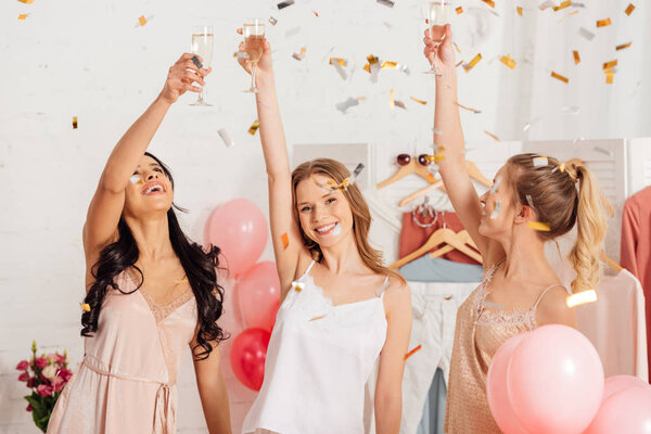 beautiful multicultural girls toasting with champagne glasses and celebrating under falling confetti during pajama party