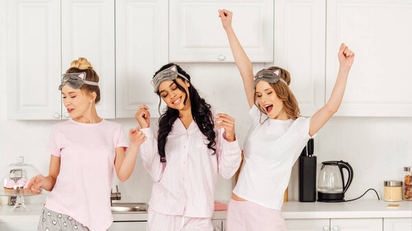 beautiful multicultural girls in sleeping masks dancing and having fun in kitchen during pajama party