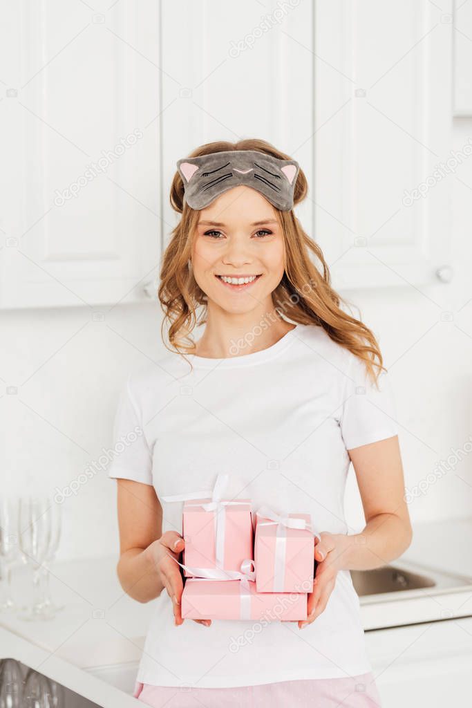 beautiful girl in pajamas and sleeping mask holding gift boxes and looking at camera in kitchen