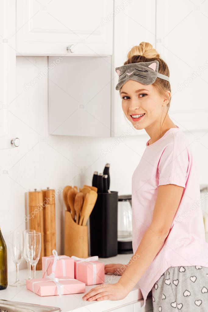 beautiful girl in pajamas and sleeping mask with pink gift boxes looking at camera in kitchen