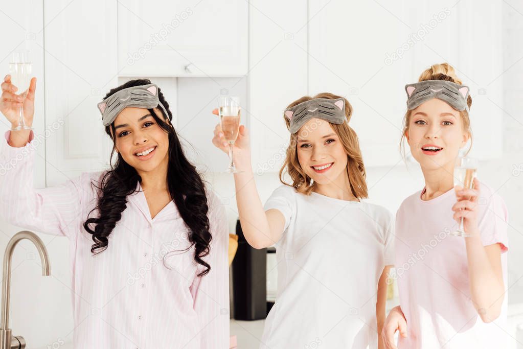 beautiful multicultural girls in sleeping masks celebrating with champagne and looking at camera during pajama party