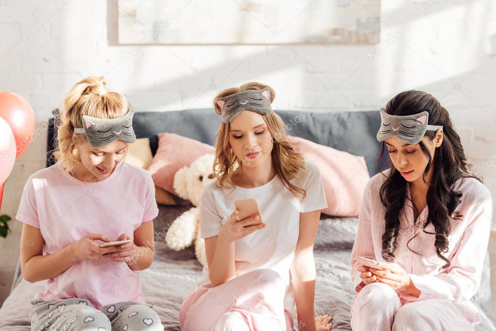 beautiful multicultural girls in sleeping masks sitting on bed and using smartphones during pajama party