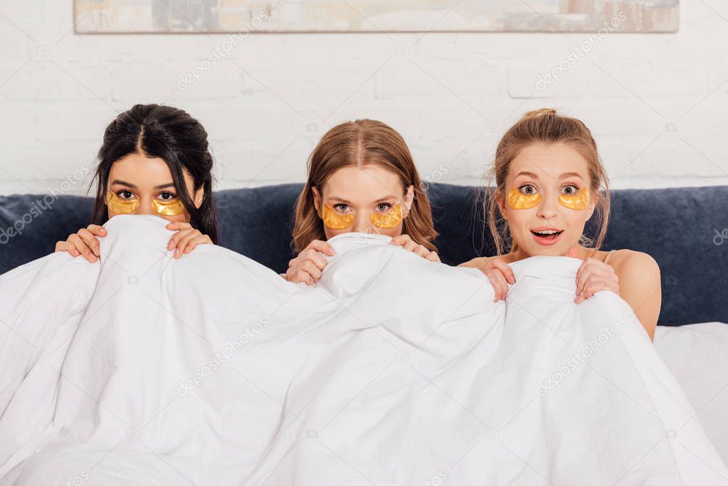 beautiful multicultural girls with golden eye patches looking at camera in bed during pajama party