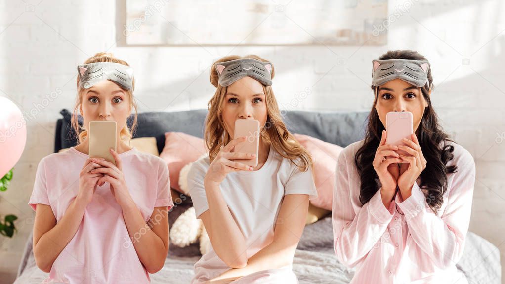beautiful multicultural girls in sleeping masks sitting on bed and covering mouths with smartphones during pajama party
