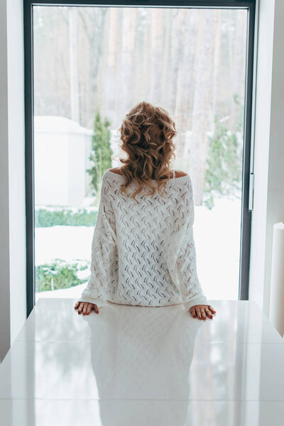 Back view of curly woman in knitted sweater looking at window