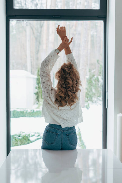 Back view of curly woman standing with hands up in front of window