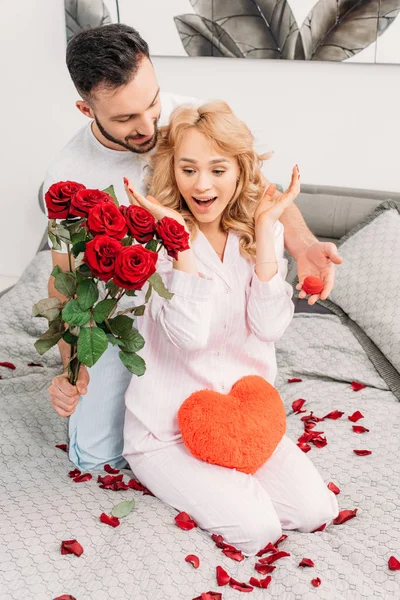 褐发女郎男子呈现鲜花和戒指盒惊讶的女朋友在卧室 — 图库照片