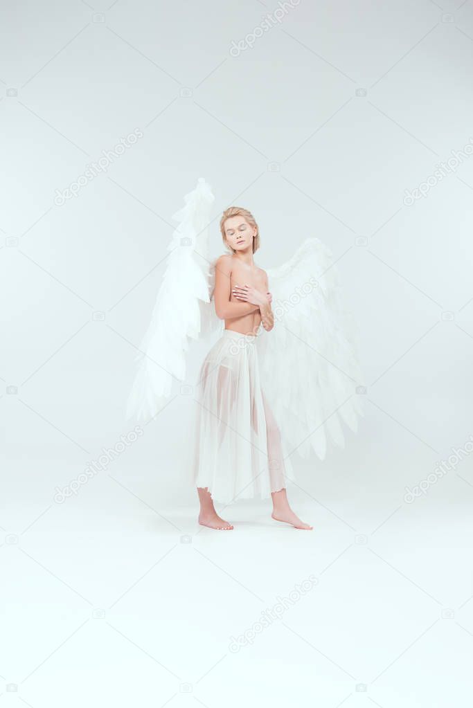 beautiful woman with angel wings covering breasts and posing with eyes closed isolated on white
