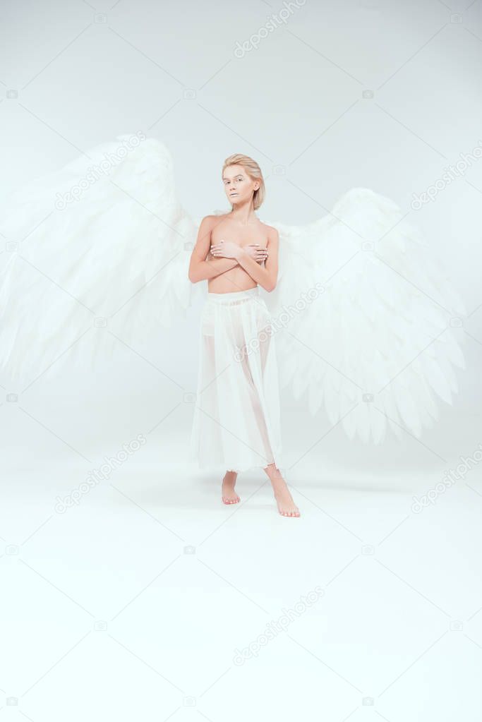 beautiful woman with angel wings covering breasts and posing while looking at camera on white background