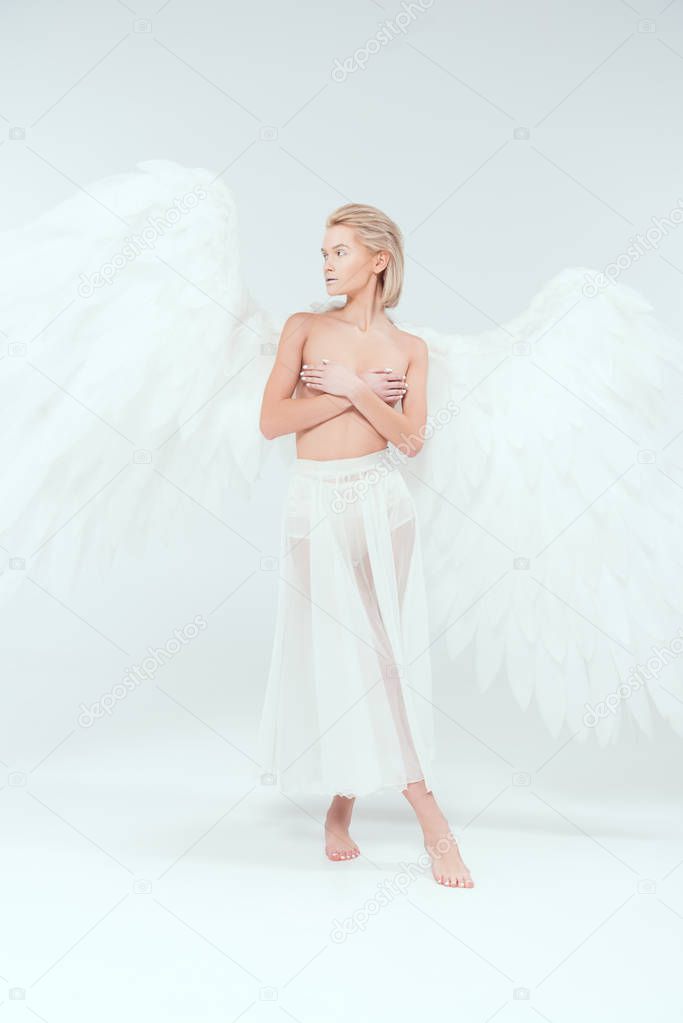 beautiful tender woman with angel wings covering breasts while looking away on white background
