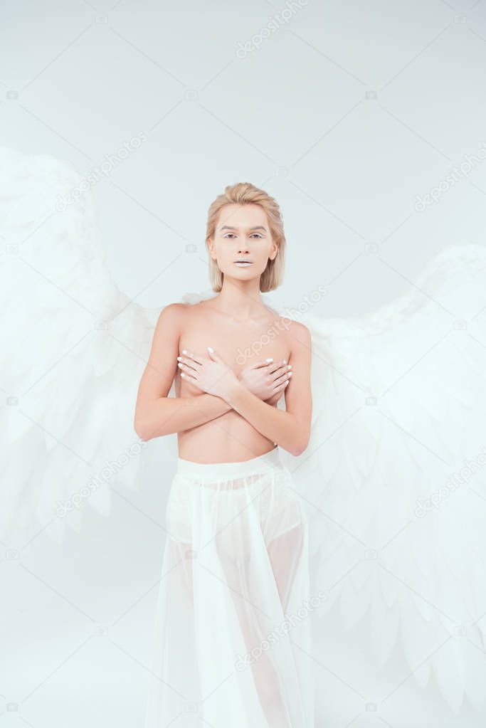 beautiful woman with angel wings covering breasts and looking at camera isolated on white