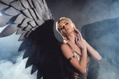 Krásná sexy žena s černým andělská křídla a zlaté doplňky dotýkání obličeje a Pózování na tmavé kouřové pozadí