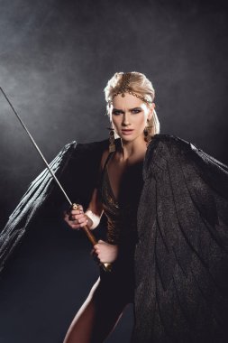 beautiful aggressive woman in warrior costume with angel wings holding sword and looking at camera on black background clipart